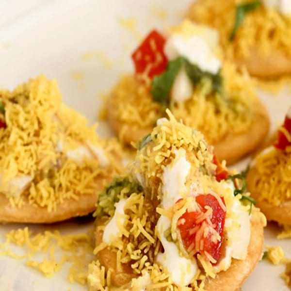 Close-up of several Indian chaat dishes, topped with yogurt, red sauce, and crispy yellow noodles.