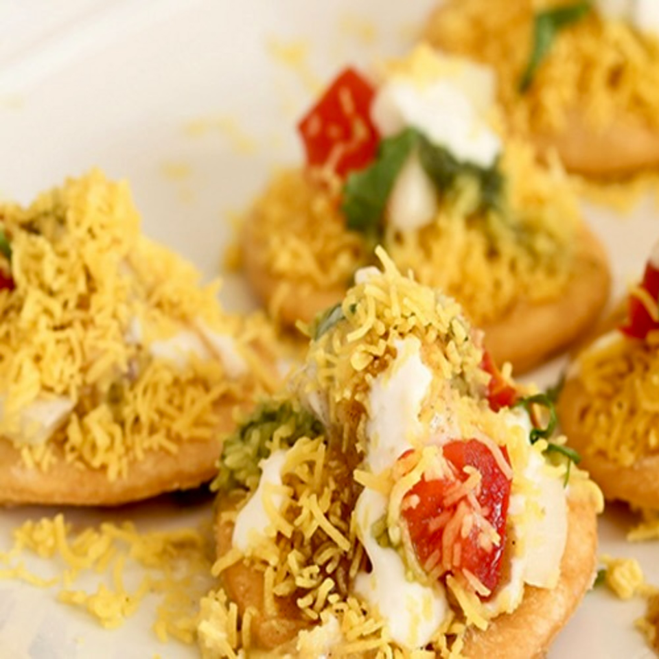 Close-up of several Indian chaat dishes, topped with yogurt, red sauce, and crispy yellow noodles.
