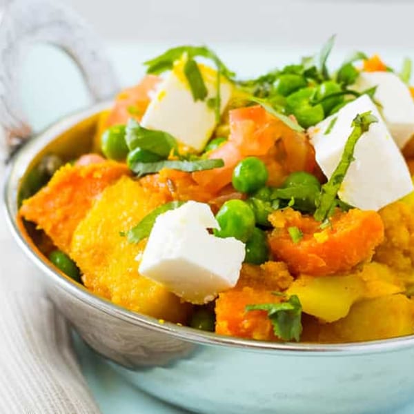 A close-up of a silver bowl filled with colorful Indian curry, topped with paneer, peas, and herbs.