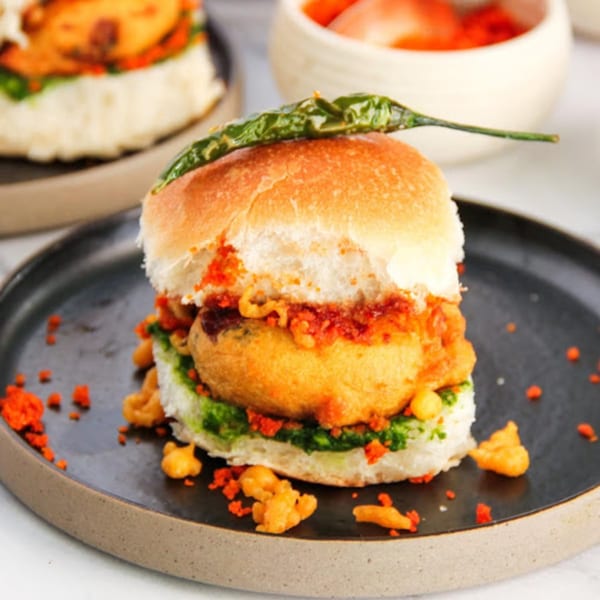 A vada pav, an Indian street food, on a dark plate, topped with a green chili.