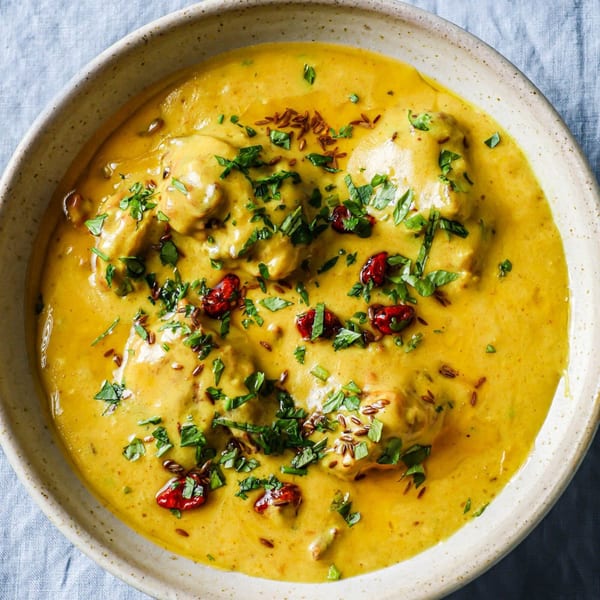 Close-up of a bowl of creamy yellow Indian curry, garnished with green herbs and red spices.