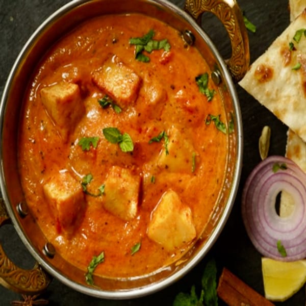 Indian paneer butter masala in a brass bowl, garnished with cilantro, served with naan, onion, and lemon.