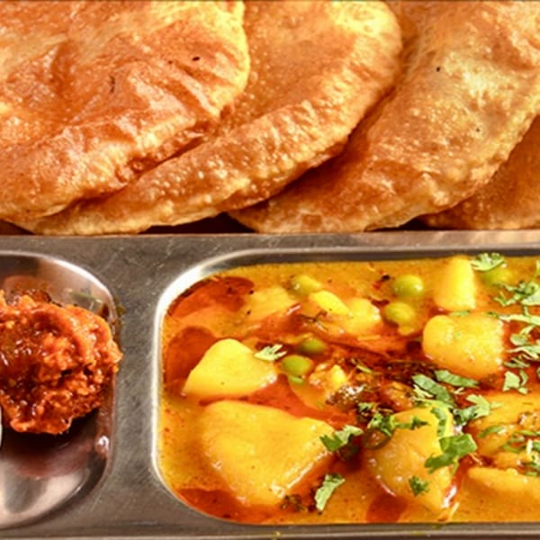 A metal tray with golden fried puri bread, potato and pea curry, and a red chutney.