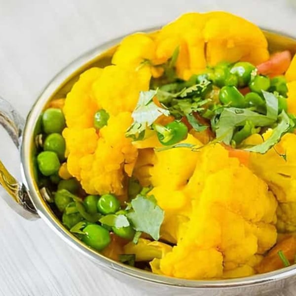 A close-up of a vibrant yellow Indian cauliflower and peas curry, garnished with fresh cilantro in a silver bowl.
