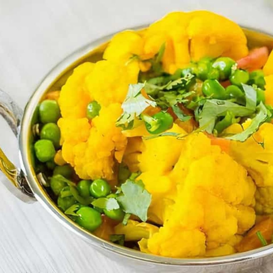 A close-up of a vibrant yellow Indian cauliflower and peas curry, garnished with fresh cilantro in a silver bowl.